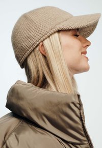 Cappello lavorato a maglia beige con visiera curva e texture a coste, abbinato a un giubbotto imbottito marrone con colletto alto e dettagli con zip.