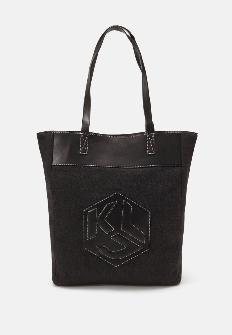 Bolso tote negro con un cuerpo de tela texturizada y acentos de cuero sintético suave. Incluye un logo hexagonal negro con costuras blancas.
