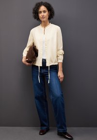 Giacca leggera color crema con colletto arricciato, maniche elastiche e lacci frontali. Abbinata a jeans blu scuri e una borsa a spalla marrone.