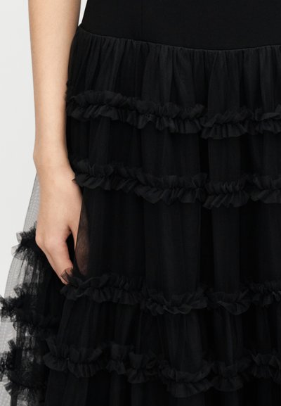 Μαύρο φόρεμα με στρώσεις από τούλι με βολάν, ημιδιαφανής υφή, στενή κορυφή και ρέουσα σιλουέτα. Χέρι κρατάει το ύφασμα.