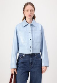 Giacca in pelle lucida, di colore blu chiaro, tagliata, con colletto, tasche frontali e bottoni a pressione, abbinata a jeans blu scuro.