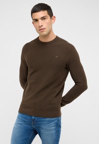 Pull en maille marron à col rond, avec des manches longues ajustées et un petit logo sur la poitrine, associé à un jean bleu.