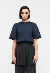 Γυναίκα που φοράει μπλε σκούρο κοντομάνικο τοπ με κυματιστά τελειώματα και σκουρόχρωμη πλισέ φούστα πάνω από αντίστοιχο σκουρόχρωμο παντελόνι με φαρδιά πόδια.