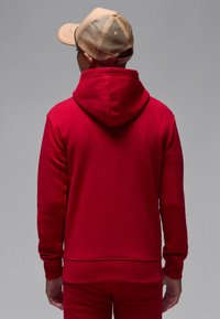 Red hoodie with a hood and front kangaroo pocket, textured fabric, ribbed cuffs, and hem. Worn with a beige mesh cap.