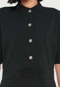 Ρούχο από μαύρο ύφασμα με ψηλό γιακά, κοντά μανίκια και μπροστινό μέρος με κουμπιά σε ασημί χρώμα. Λείος υφής, εφαρμοστό σχέδιο.