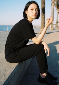 Mlada žena u crnoj odjeći sjedi na betonskoj ivičnjaku kraj palmi i šetnice uz ocean danju, gledajući prema kameri.