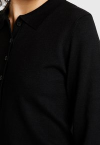 Zwart gebreid poloshirt met kraag, voorzien van een knoopsluiting en een geribbelde structuur, gefocust op een close-up van het bovenlichaam.