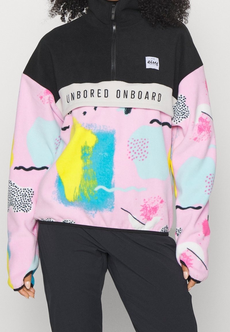 Veste polaire noir et rose à blocs de couleurs avec des motifs abstraits et le texte "UNBORED ONBOARD" sur la poitrine, portée avec un pantalon noir.