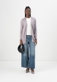 Helllila Strickjacke, weißes Hemd, weit geschnittene blaue Jeans, grau-beige Schuhe, schwarze Handtasche, glattes Material, lässige Passform.