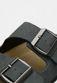 Šedá semišová sandál s dvěma nastavitelnými popruhy a metalickými přezkami. Interiér je v béžové barvě a ukazuje měkkou, strukturovanou stélku.