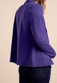 Fioletowa kurtka z długim rękawem o luźnym kroju, ze sztywnym kołnierzem i kieszeniami bocznymi. Gładka tekstura, minimalne szwy i lekko zaokrąglony dół.