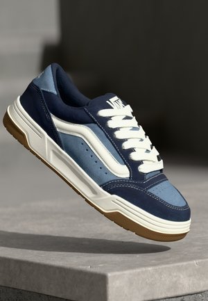 Sinine ja tumesinine sneaker, mis on valmistatud sametist ja nahast, valgete detailide, kummitaldade, õmmeldud elementide ja paeltega sulgemisega.