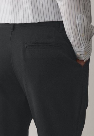 Bărbat purtând pantaloni eleganți gri închis și o cămașă albă cu dungi verticale gri, mână în buzunar, văzut de la talie în jos.