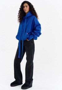 Hanorac albastru supradimensionat cu buzunar kangaroo, șnururi și accente roz, asortat cu blugi negri cu picior drept și pantofi negri.
