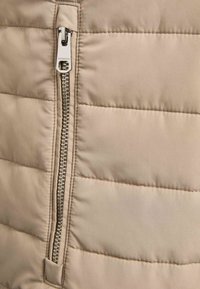 Chaleco acolchado beige con diseño acolchado vertical, cremallera metálica plateada y textura suave. La cremallera corre a lo largo del frente.