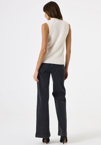 Vrouw staand met de rug naar de kijker, draagt een mouwloos wit textuurtopje, zwarte wijde jeans met hoge taille en zwarte hakken.