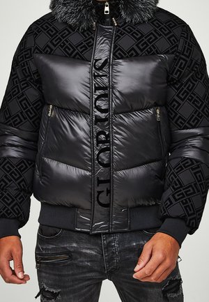 Doudoune noire à motifs avec col en fourrure et grand panneau matelassé central brillant avec texte vertical noir, portée avec un jean noir déchiré.