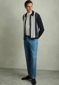Cardigan cu dungi bleu marin și alb, purtat cu un tricou alb simplu, blugi drept de culoare albastră și mocasini negri. Textură netedă.