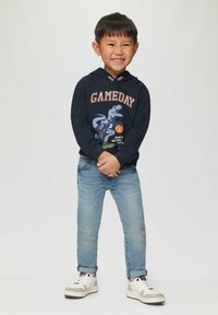 Marineblauwe hoodie met dinosaurusafbeelding en de tekst "GAMEDAY", gecombineerd met lichtblauwe spijkerbroek en witte sneakers met grijze accenten. Kind glimlachend, met gekruiste armen.