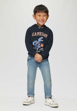 Marineblauwe hoodie met dinosaurusafbeelding en de tekst "GAMEDAY", gecombineerd met lichtblauwe spijkerbroek en witte sneakers met grijze accenten. Kind glimlachend, met gekruiste armen.
