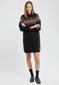 Robe noire en tricot avec un yoke à motifs présentant des designs géométriques rouges et crème. Comprend des manches longues et un petit logo doré en bas.