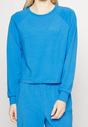 Person iført en knaldblå langærmet top og matchende bukser med subtile tekst detaljer på brystet og låret.