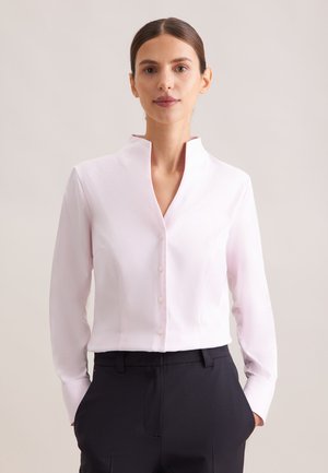 Femme portant une chemise rose clair boutonnée avec un col montant et un pantalon noir, debout les mains dans les poches devant un fond uni.