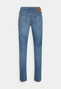 Jean en denim bleu avec couture classique, deux poches arrière, un patch en cuir marron sur la ceinture et une étiquette en tissu rouge sur la poche droite.