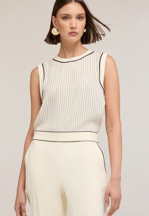 Kvinde med kort brunt hår iført ærmeløs hvid top med tynde striber og matchende højtaljede bukser med store guld- og hvide øreringe.