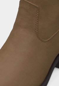 Κοντινό πλάνο καφέ δερμάτινου μποτάκι αστραγάλου που δείχνει την ομαλή υφή, τις λεπτομέρειες ραφής και μέρος από χαμηλό μαύρο τακούνι.