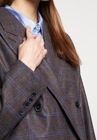 Osoba upravující límec světle modré košile s knoflíky, oblečená v hnědém kostkovaném saku s tmavými knoflíky, s dlouhými hnědými vlasy viditelnými.