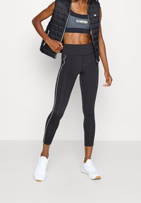 Černá zateplená vesta na zip, kombinovaná s tmavě modrou sportovní podprsenkou se zlatým textem, černými legínami s bílými bočními proužky a bílými teniskami.