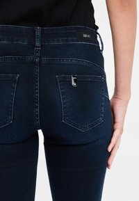 Jeans skinny blu scuro con un design elegante, caratterizzati da un accento metallico sulla tasca posteriore e un'etichetta con il marchio in vita.