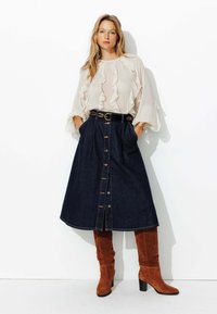 Jupe en denim avec un design à boutons sur le devant, une taille cintrée et des poches latérales, associée à un chemisier léger à volants et des bottes hautes en daim marron.