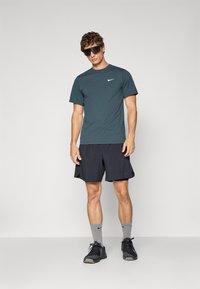 Mørk tealfarget Nike t-skjorte, svarte shorts, grå treningssokker og svarte sko. Modellen står mot en ensfarget bakgrunn iført svarte solbriller.