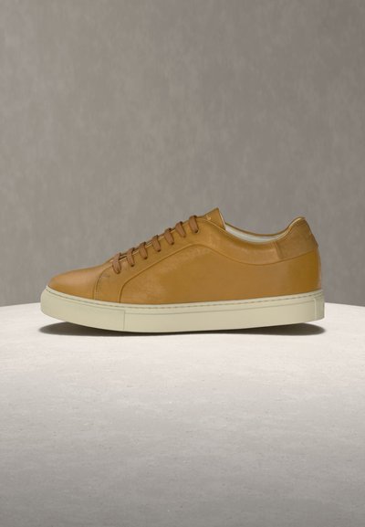 Ταμπά δερμάτινο sneaker χαμηλού κοψίματος με καφέ κορδόνια και λευκή ελαστική σόλα σε ελαφρώς υφή επιφάνεια πάνω σε ουδέτερο φόντο.