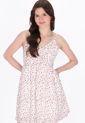 Młoda kobieta z długimi brązowymi włosami, ubrana w białą sukienkę w kwiaty i czerwone kolczyki w kształcie wiśni, stoi z rękami na biodrach.