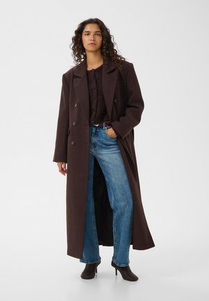 Kvinde iført en lang mørkebrun frakke, mørk sweater, blå jeans og sorte ankelstøvler med hæl, står med den ene hånd i lommen.