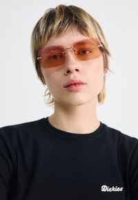 Rechteckige Sonnenbrillen mit orange getönten Gläsern, dünnen goldenen metallischen Rahmen und einem schwarzen T-Shirt mit weißen gestickten Text.