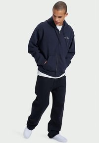 Navy blue zip-up hoodie made of soft fabric, featuring a kangaroo pocket and drawstring hood, worn over a white T-shirt and dark pants.
