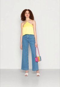 Top amarelo sem mangas combinado com calças de ganga azuis de cintura alta e perna larga. Os acessórios incluem uma bolsa multicolorida e sandálias brancas.