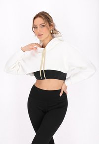 Witte cropped hoodie met gouden koorden, gecombineerd met een zwarte sportbeha en hoge zwarte leggings. Gladde stof, relaxte pasvorm.