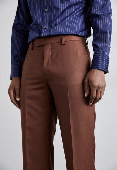 Pantalon marron ajusté en tissu lisse, avec une coupe droite et des poches latérales, associé à une chemise rayée bleu foncé.