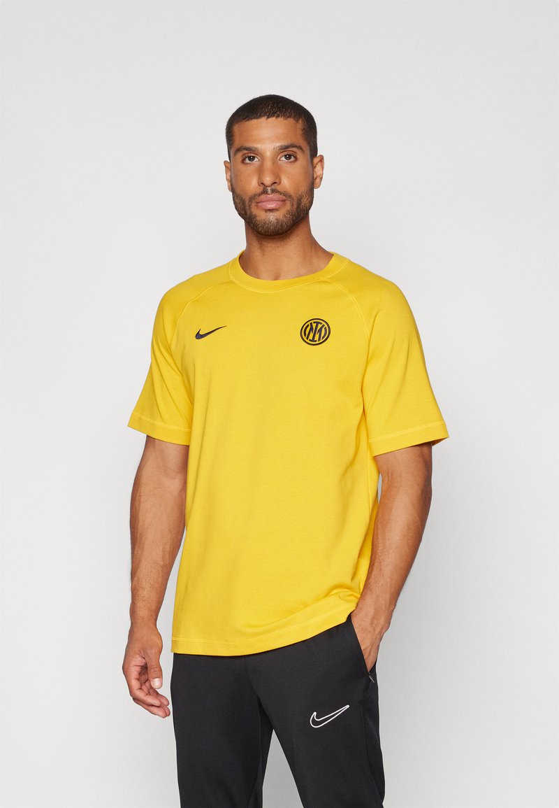 Mand iført en gul Nike T-shirt med et cirkulært sort og gul emblem og sorte Nike bukser, stående mod en ensfarvet baggrund.