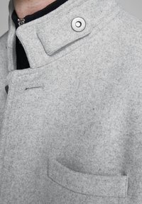 Nahaufnahme eines hellgrauen Wollmantels mit hohem Kragen, einem metallischen Druckknopf und einer horizontalen Tasche im Brustbereich.