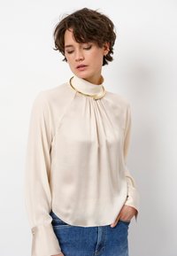 Crème-kleurige zijden blouse met een hoge kraag, plooien aan de voorkant en lange mouwen. Bevat gouden hardware accenten bij de halslijn.