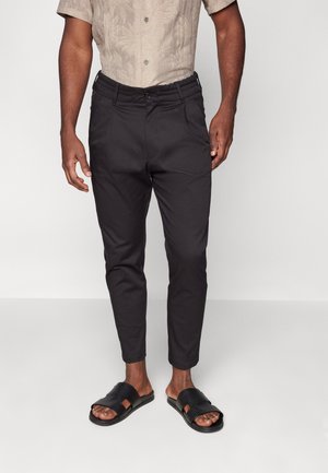 Mann trägt schwarze, maßgeschneiderte, knöchellange Hose, beige Kurzarmhemd und schwarze offene Zehensandalen, steht vor weißem Hintergrund.
