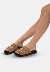 Chaussons en daim brun avec deux accents de boucles en argent réglables, dotés d'une semelle en caoutchouc noir rembourrée et d'un design ouvert à l'avant.