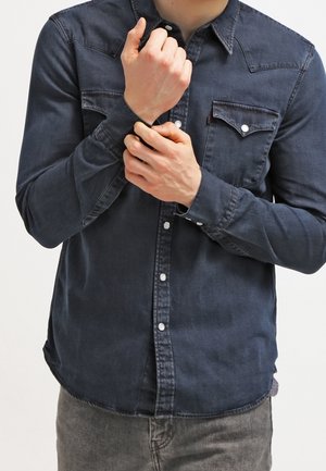 Mann mit dunklem Jeanshemd, der den Manschettenknopf seines linken Handgelenks schließt, vor einem schlichten hellen Hintergrund.