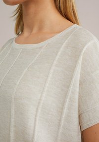 Gaiši beiga adīta tops ar apaļu kakla izgriezumu, īsām piedurknēm un vertikālām tekstūrām, ar smalku spīdumu visā virsmā.